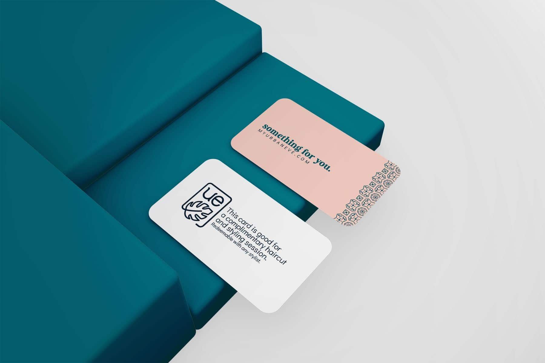 Two business cards placed on teal rectangular blocks. The upper card is pink and reads 