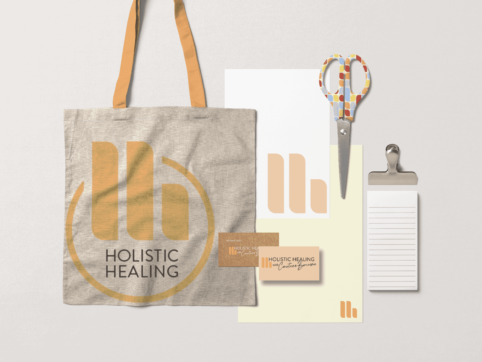 A collection of branded items for Holistic Healing is shown, including a beige tote bag, a pair of scissors with a colorful handle, a business card, a letterhead, a notepad, and a clipboard. The items feature a logo with the letter 