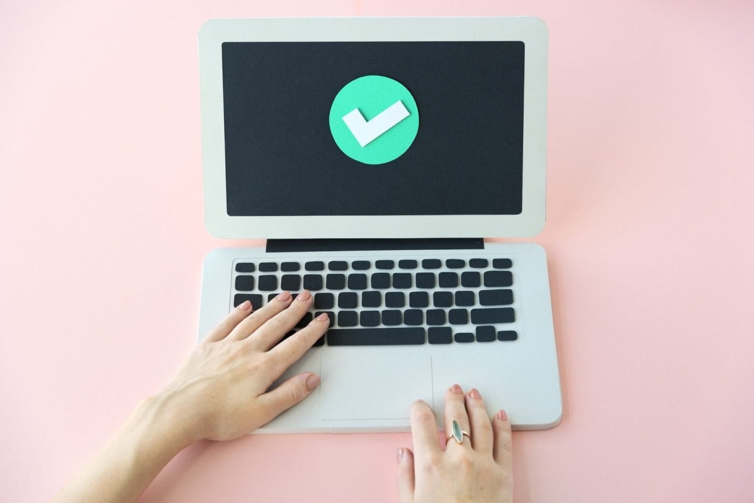 A person’s hands on a laptop keyboard with a large check mark icon—a clean web displaying vector file, such as SVG files—displayed on the screen, set against a pink background. - Market Design Team: Define. Structure. Expand.