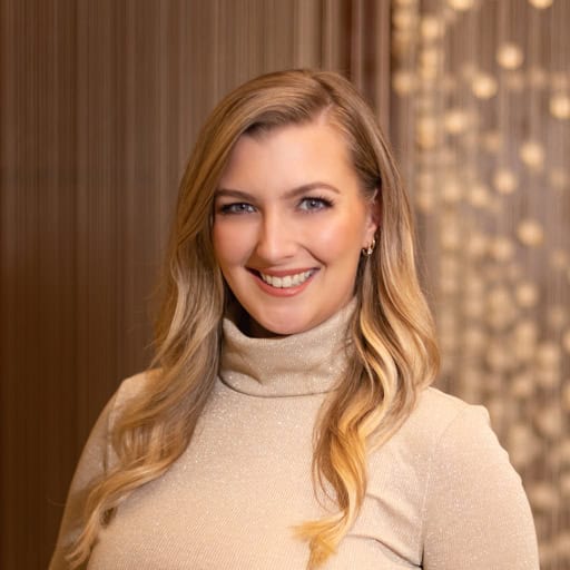 A person with long blonde hair is smiling, wearing a light-colored turtleneck sweater that matches the soft professionalism of a Winnow Financial setting. The background is softly blurred with circular lights, evoking a warm and welcoming atmosphere. - Market Design Team: Define. Structure. Expand.