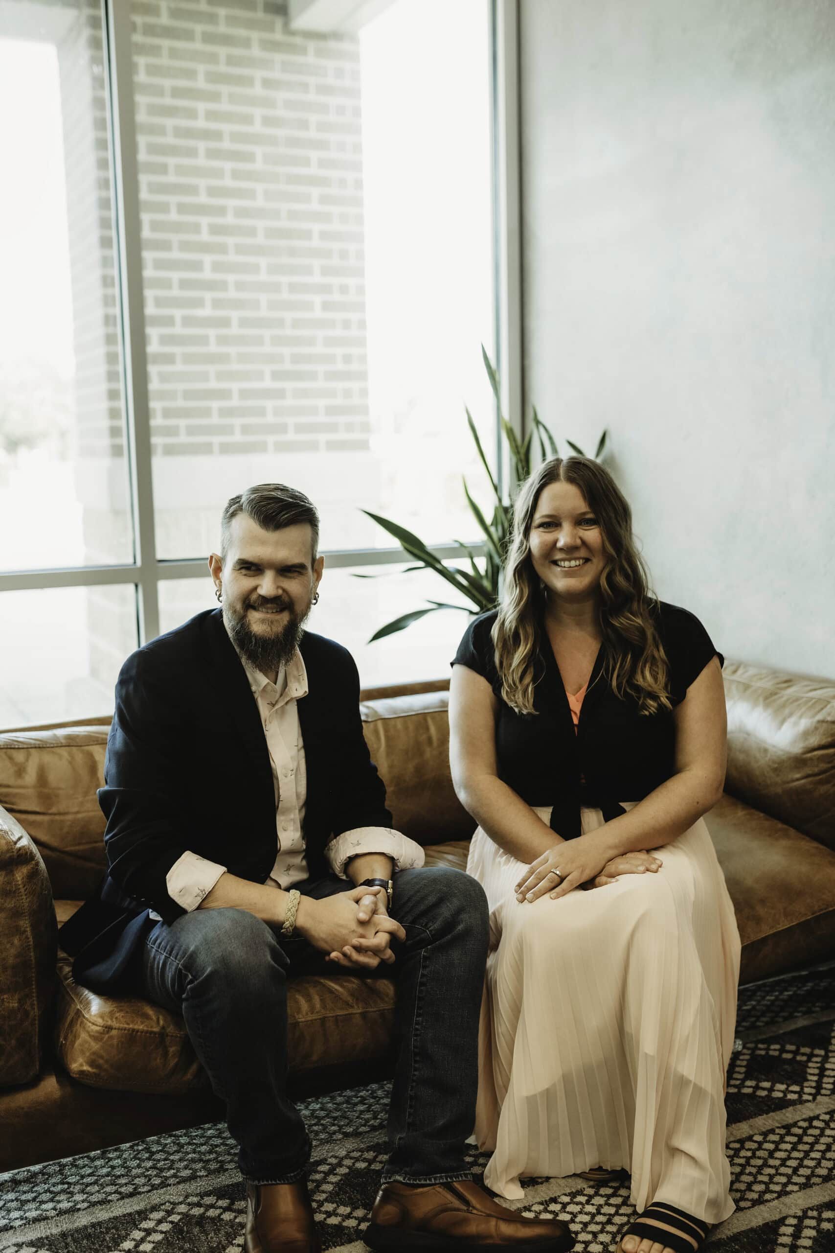 A smiling man and woman sit on a brown leather couch. The man has a beard and is wearing a dark blazer and light shirt. The woman is wearing a black top and a long white skirt. A plant is visible in the background near a large window. - Market Design Team: Define. Structure. Expand.