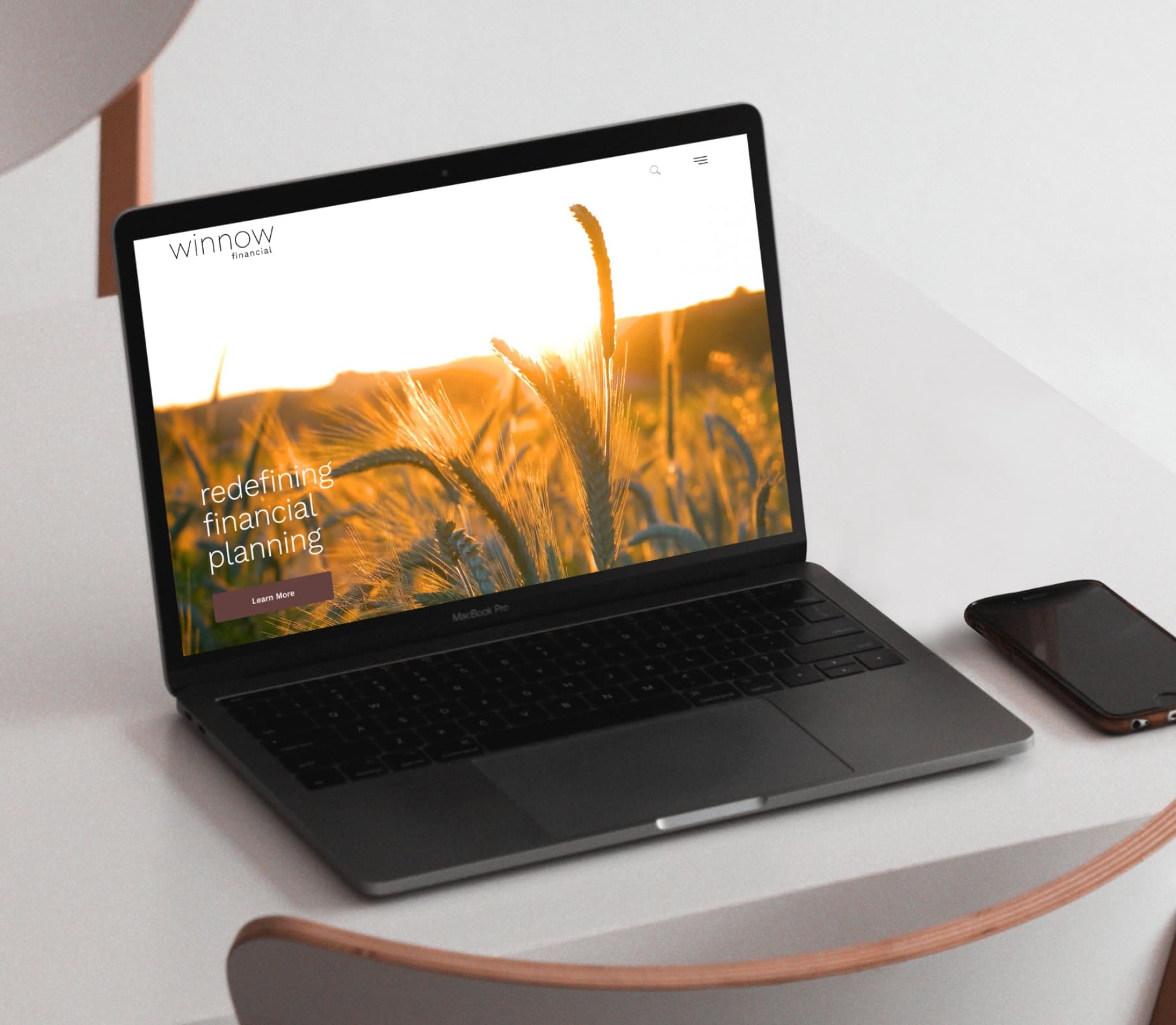A MacBook Pro sits on a white table with a phone beside it. The laptop screen displays the Winnow Financial website, featuring an image of a wheat field and the text 