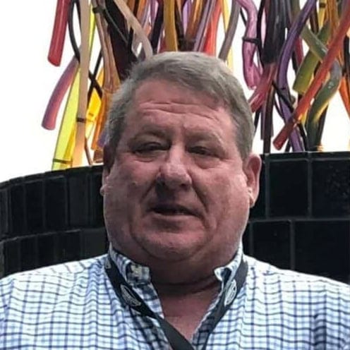 A man with short gray hair wearing a blue and white checkered shirt stands in front of a black Felder Water Well structure, with colorful vertical tubes or pipes in the background. - Market Design Team: Define. Structure. Expand.