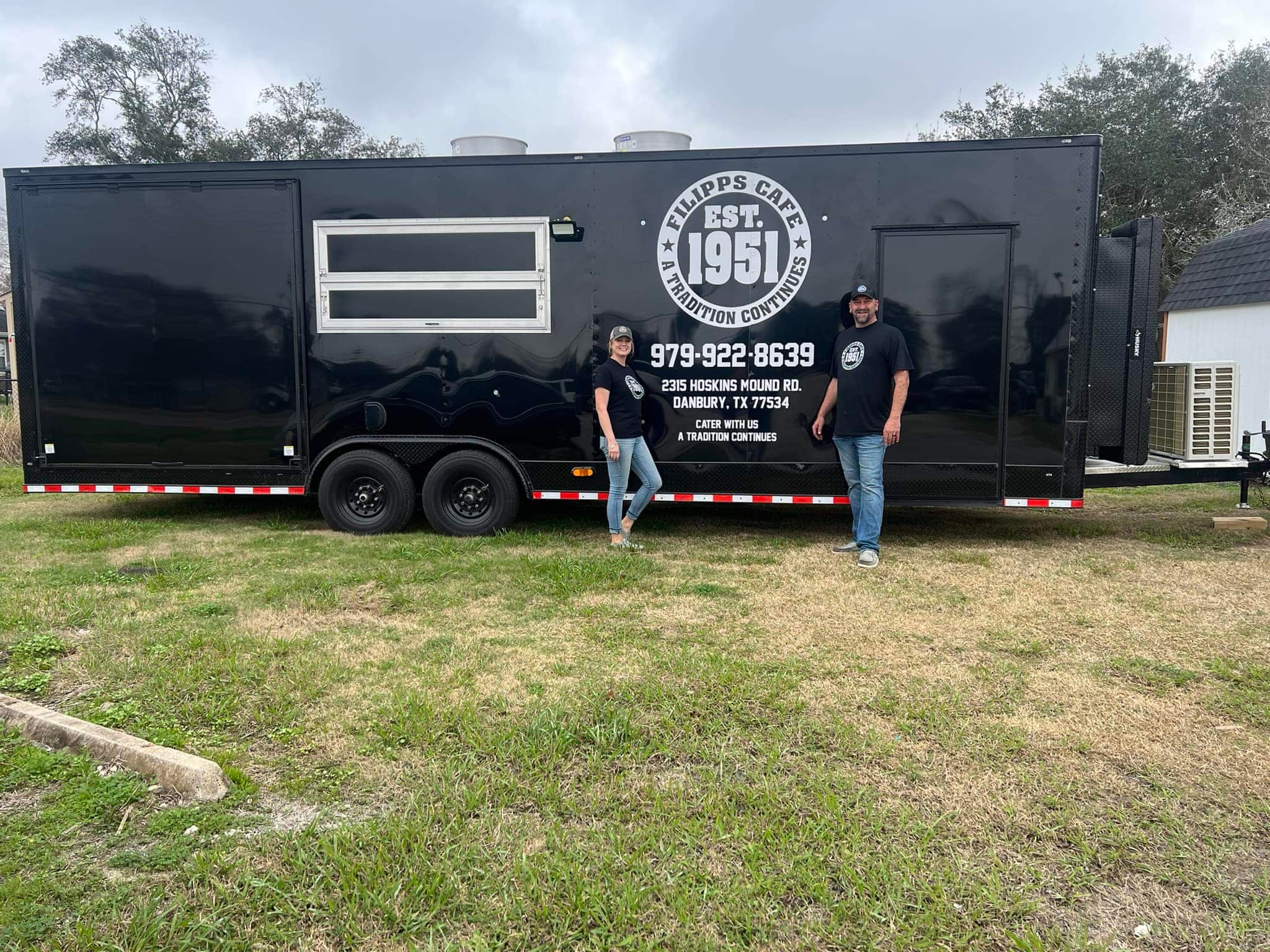 Two people stand in front of a large black Filipp's Cafe food trailer parked on grass. The trailer displays 