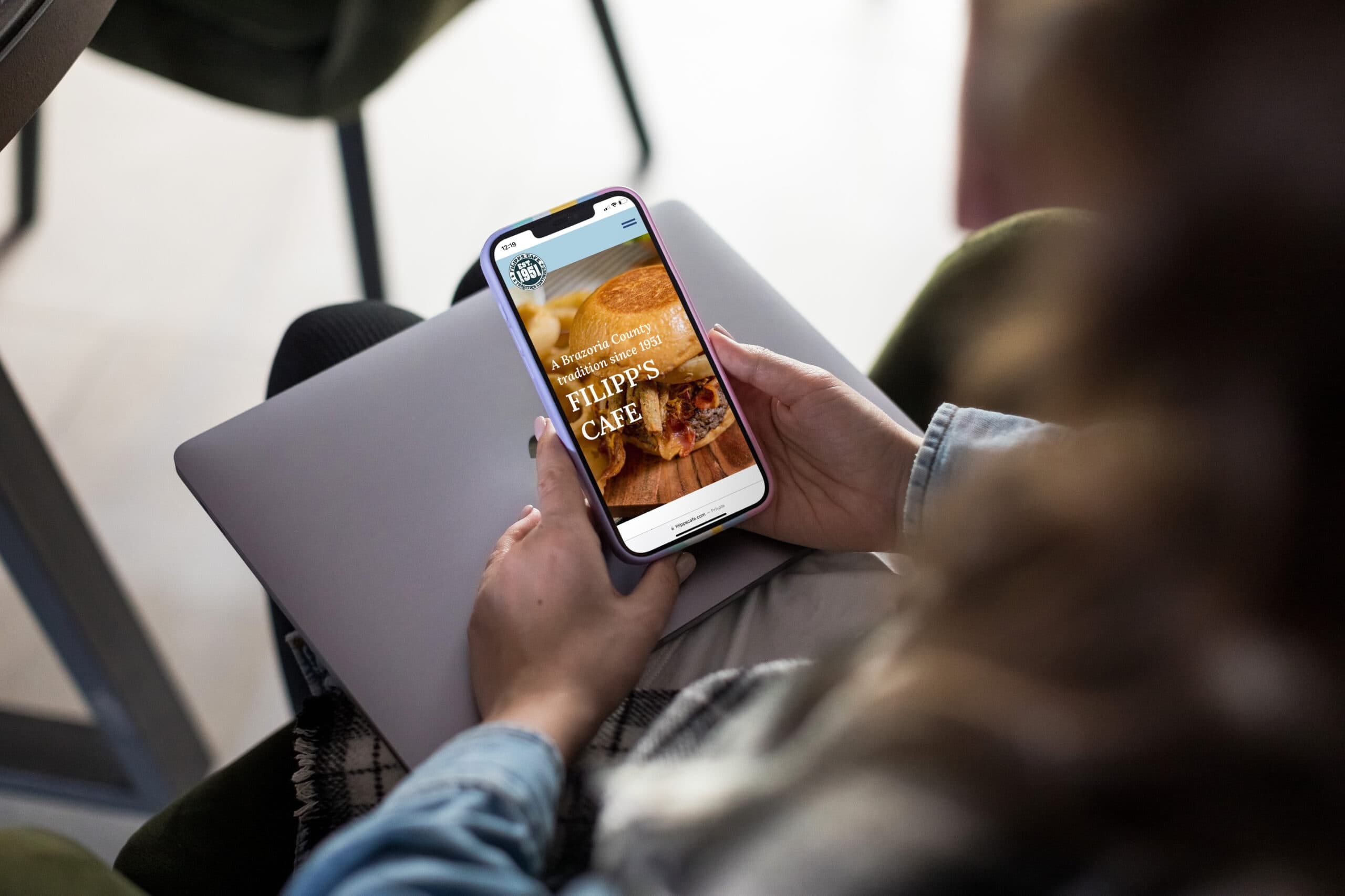 A person, partially covered by a blanket, sits with a closed laptop on their lap while holding a smartphone that displays Filipp's Cafe’s website featuring an enticing food image. - Market Design Team: Define. Structure. Expand.