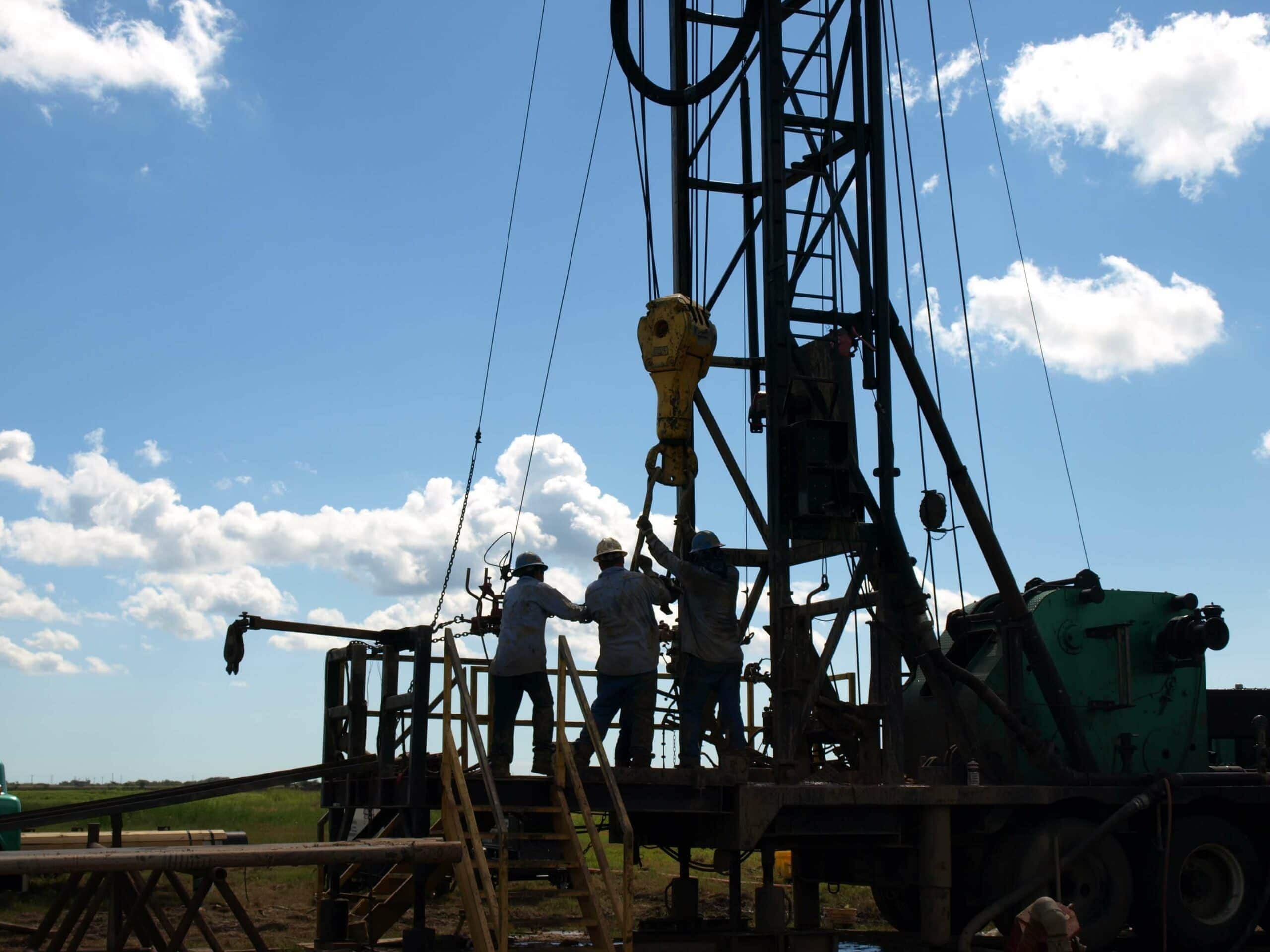 Three workers operate machinery on a Felder Water Well drilling rig outdoors during daylight, silhouetted against a blue sky with scattered clouds—a striking scene of well drilling in action. - Market Design Team: Define. Structure. Expand.