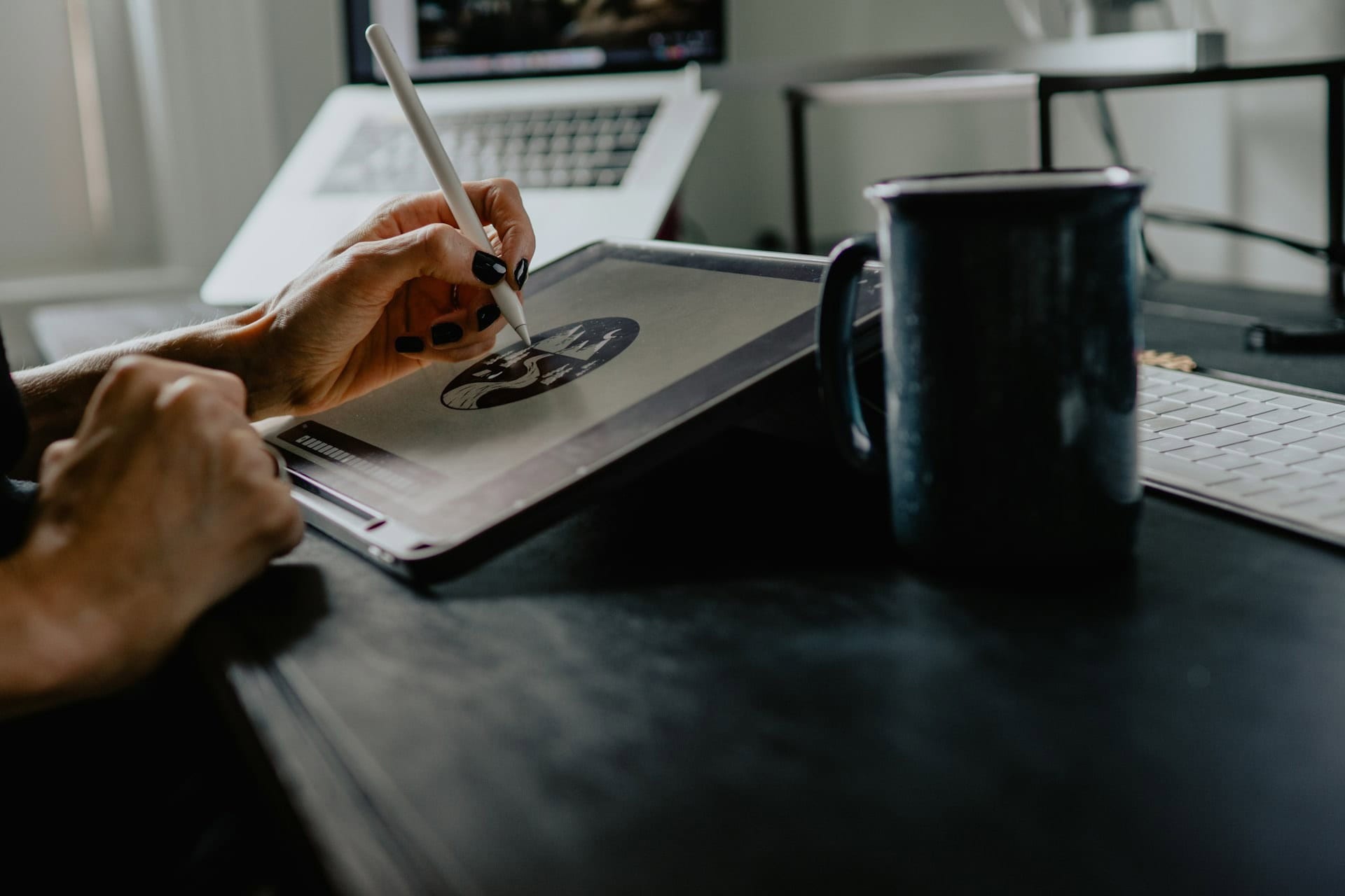 A person uses a stylus to draw on a graphics tablet at a desk, perfect for logo graphic designers crafting a professional logo. A laptop, keyboard, and dark mug sit nearby, all illuminated by natural light from a window. - Market Design Team: Define. Structure. Expand.