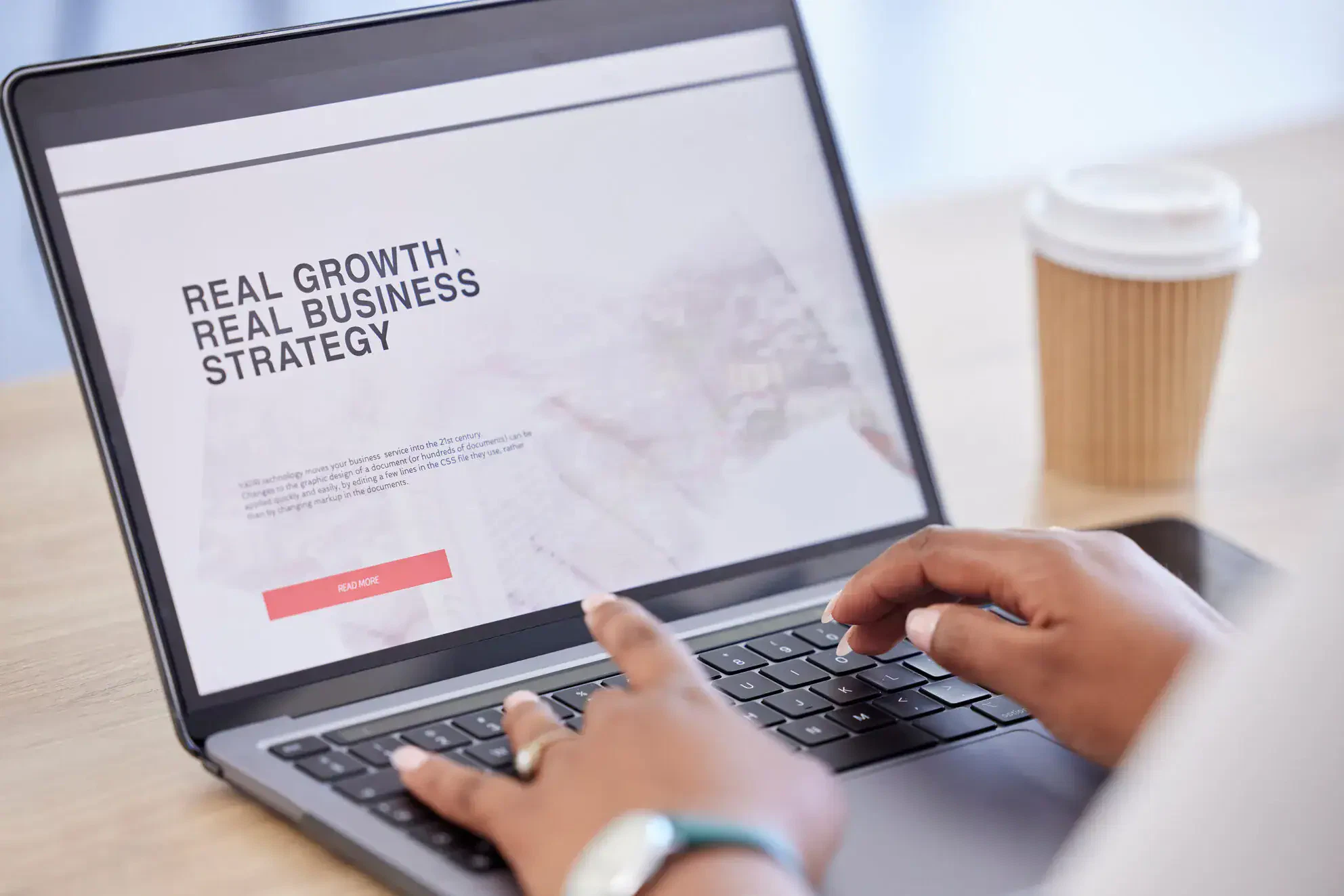 A person types on a laptop displaying a website with the text “Real Growth. Real Business Strategy,” highlighting SEO Services. A disposable coffee cup sits on the table next to the laptop. - Market Design Team: Define. Structure. Expand.