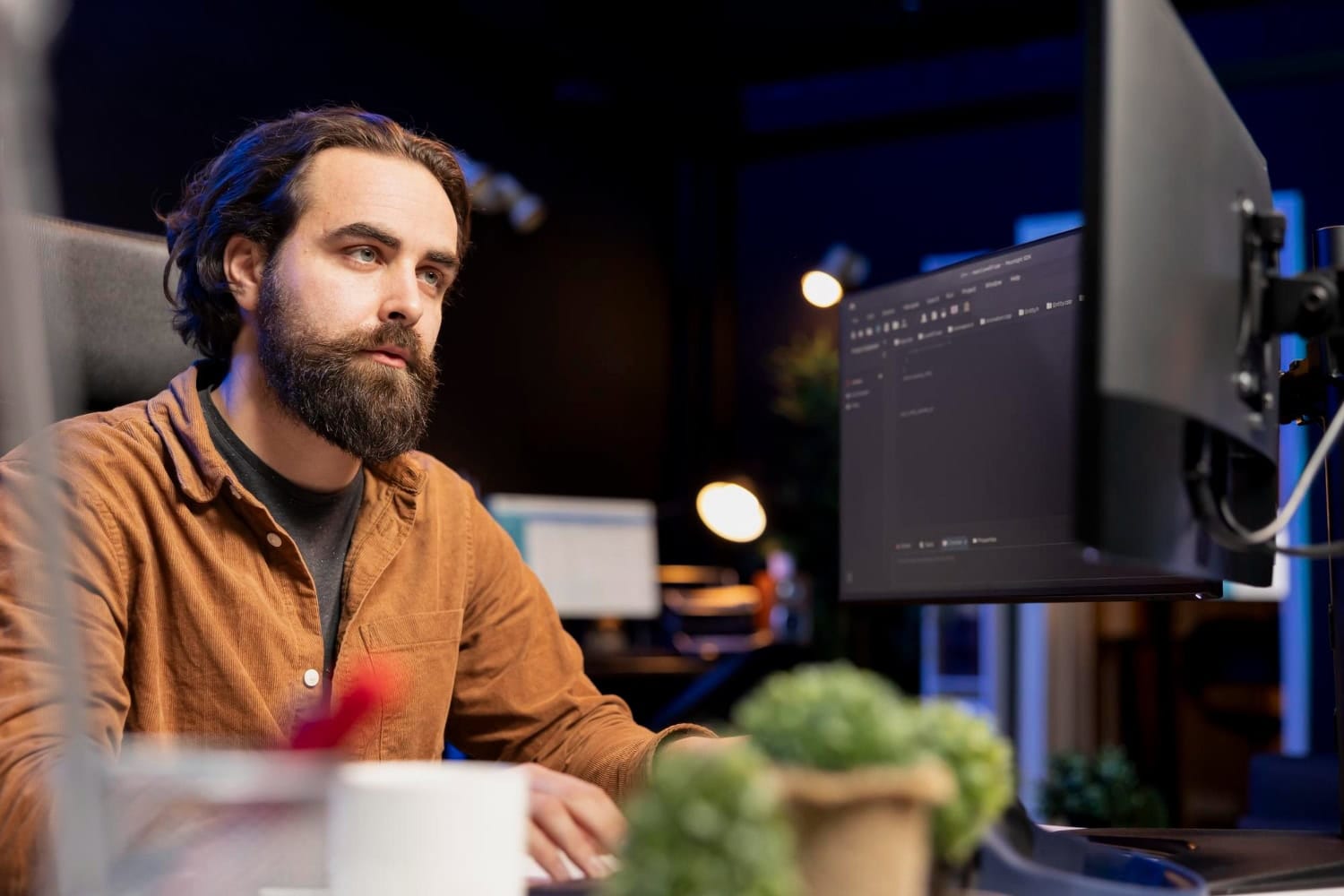 A man with a beard and long hair, wearing a brown shirt, sits at a desk and looks at a computer monitor in a dimly lit office with plants and equipment, focused on expert web development to streamline business operations. - Market Design Team: Define. Structure. Expand.