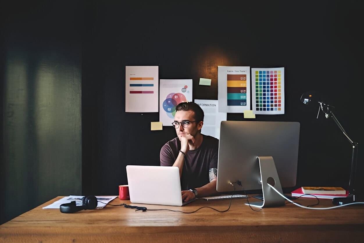 A web page designer sits at a desk with a laptop and desktop,