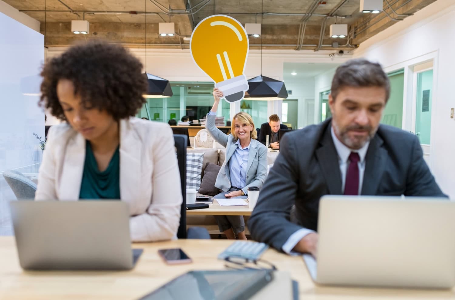 A woman in the background holds up a large paper cutout of a lightbulb and smiles, symbolizing creative solutions, while two people in the foreground work on laptops tackling business problems in an office setting. - Market Design Team: Define. Structure. Expand.