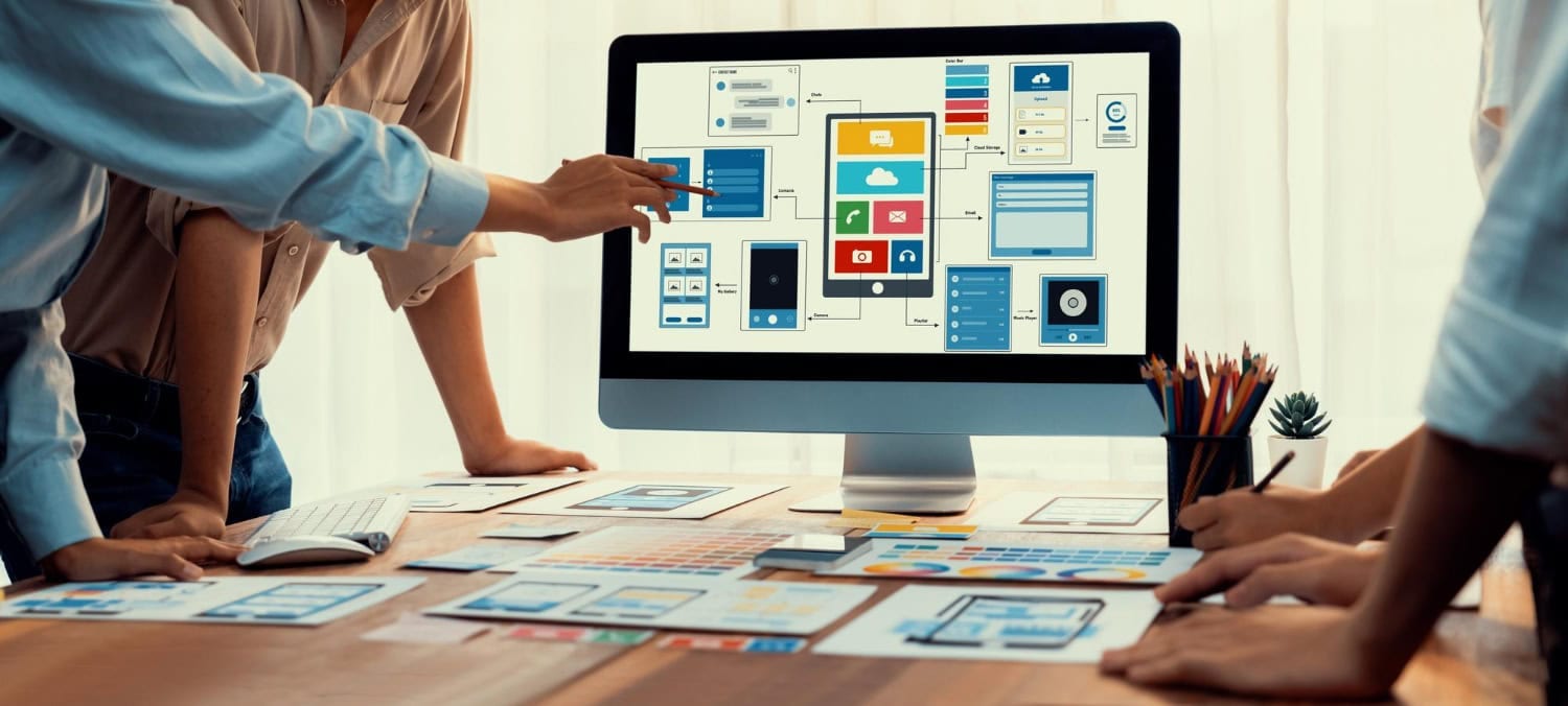 Three people stand around a desk discussing a computer screen displaying a website wireframe. Papers and color samples are spread out, suggesting local web designers collaborating on winter projects in this creative planning meeting. - Market Design Team: Define. Structure. Expand.