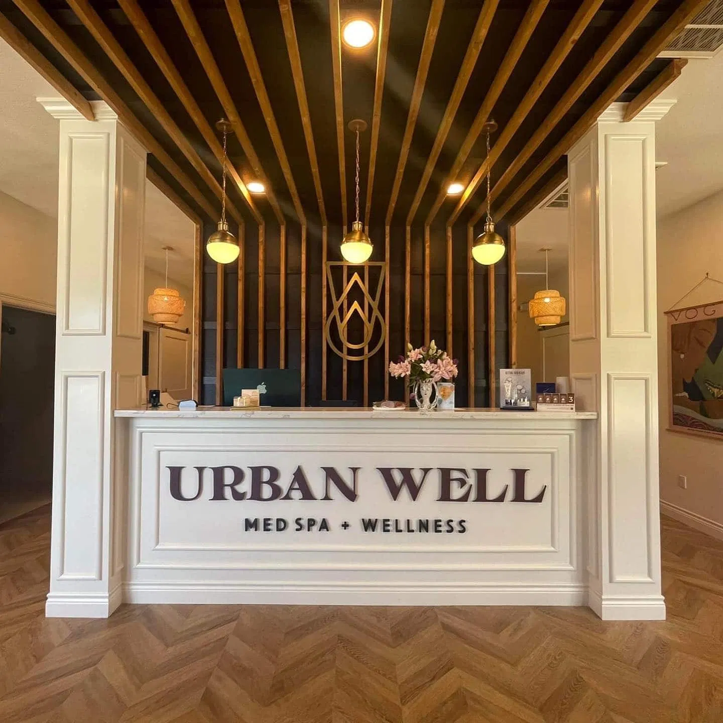 Reception area of Urban Well Med Spa + Wellness with a modern design. The front desk features the spa's name, with warm lighting and decorative elements, including a logo on the wall and floral accents on the counter. - Market Design Team: Define. Structure. Expand.