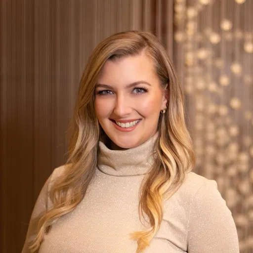 A person with long blonde hair is smiling, wearing a light-colored turtleneck sweater that matches the soft professionalism of a Winnow Financial setting. The background is softly blurred with circular lights, evoking a warm and welcoming atmosphere. - Market Design Team: Define. Structure. Expand.