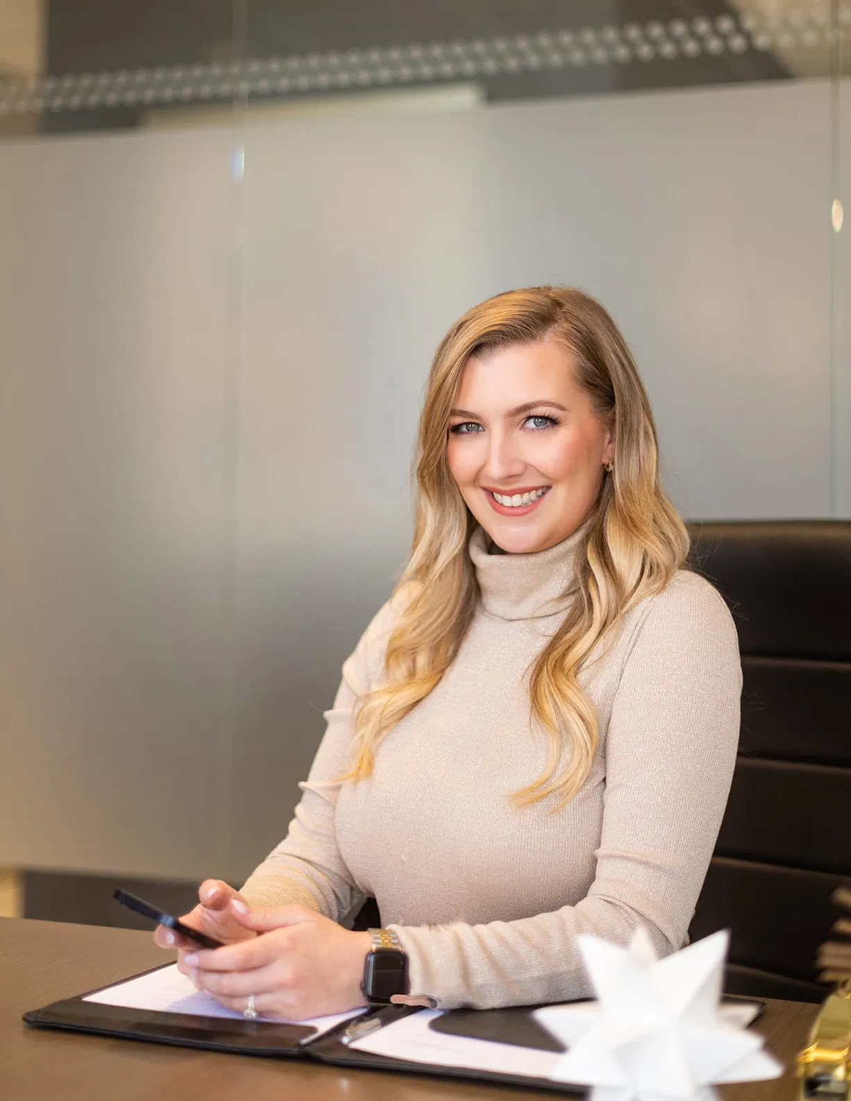 A woman with long blonde hair sits at a desk, smiling at the camera. She is holding a smartphone and wearing a light-colored turtleneck sweater, representing Winnow Financial's approachable and modern style. A closed binder rests on the desk in the professional office setting behind her. - Market Design Team: Define. Structure. Expand.