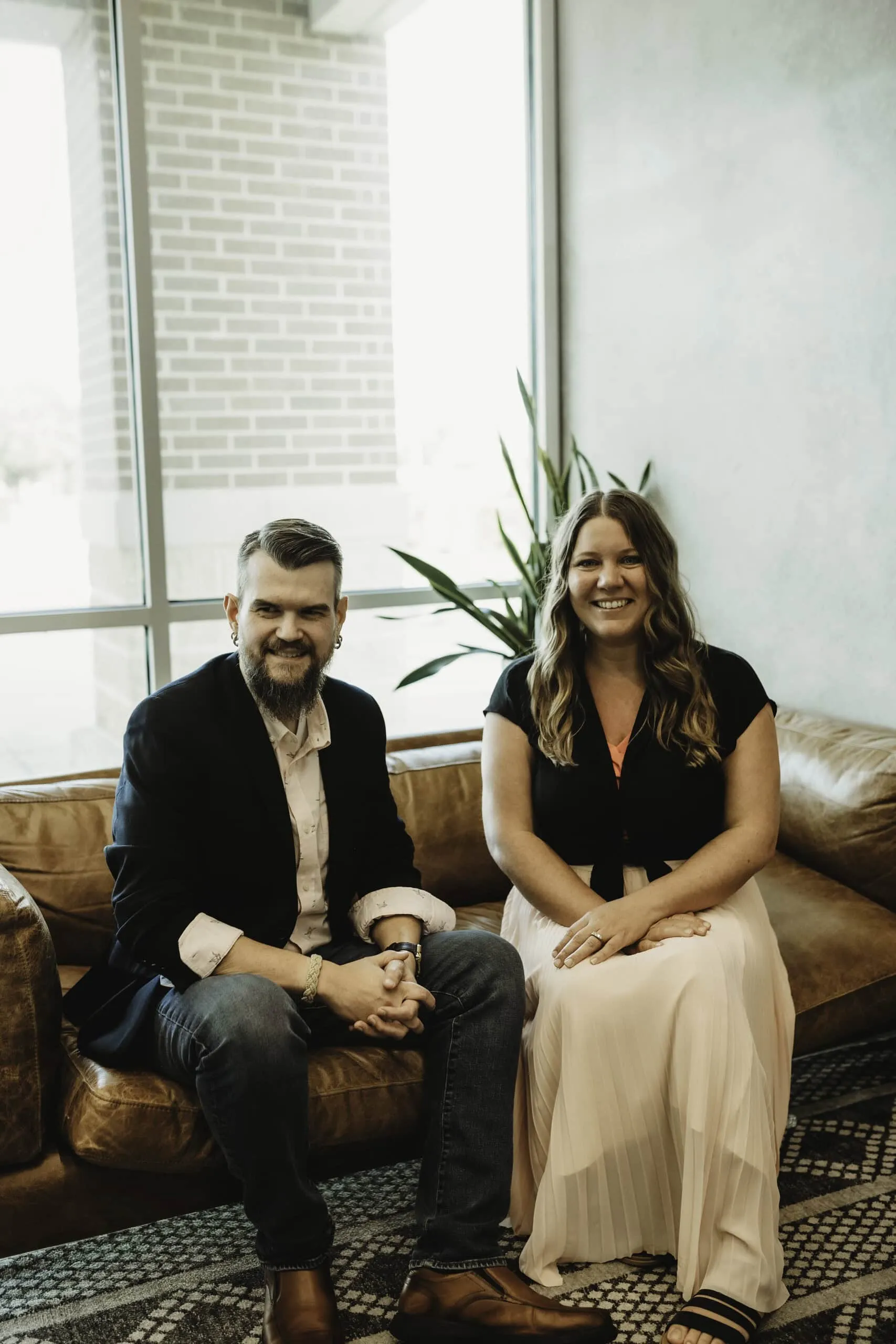A smiling man and woman sit on a brown leather couch. The man has a beard and is wearing a dark blazer and light shirt. The woman is wearing a black top and a long white skirt. A plant is visible in the background near a large window. - Market Design Team: Define. Structure. Expand.