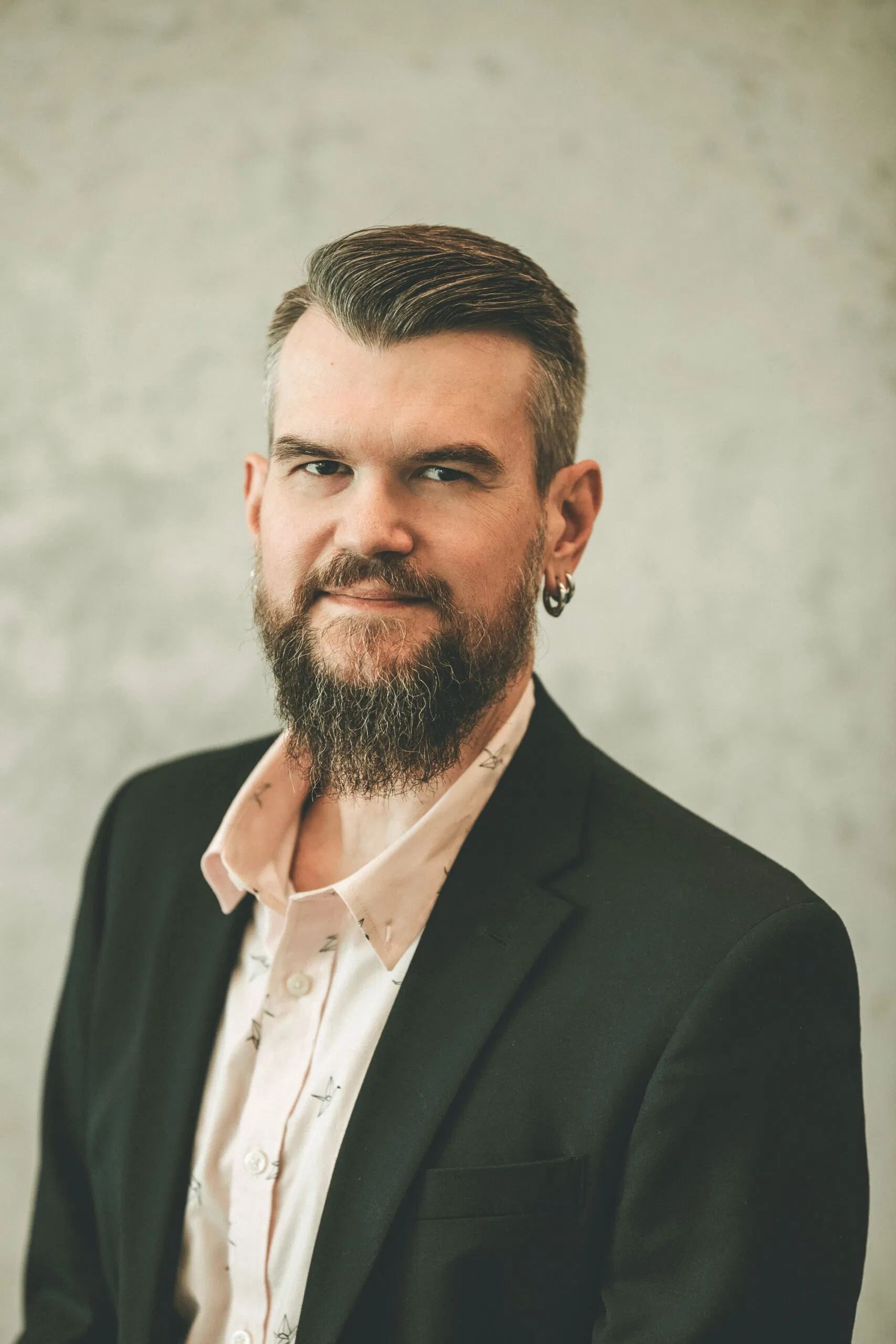 A person with a beard and short hair is wearing a black blazer over a light-colored shirt. They have an earring in the left ear. The background is a plain, neutral tone. - Market Design Team: Define. Structure. Expand.
