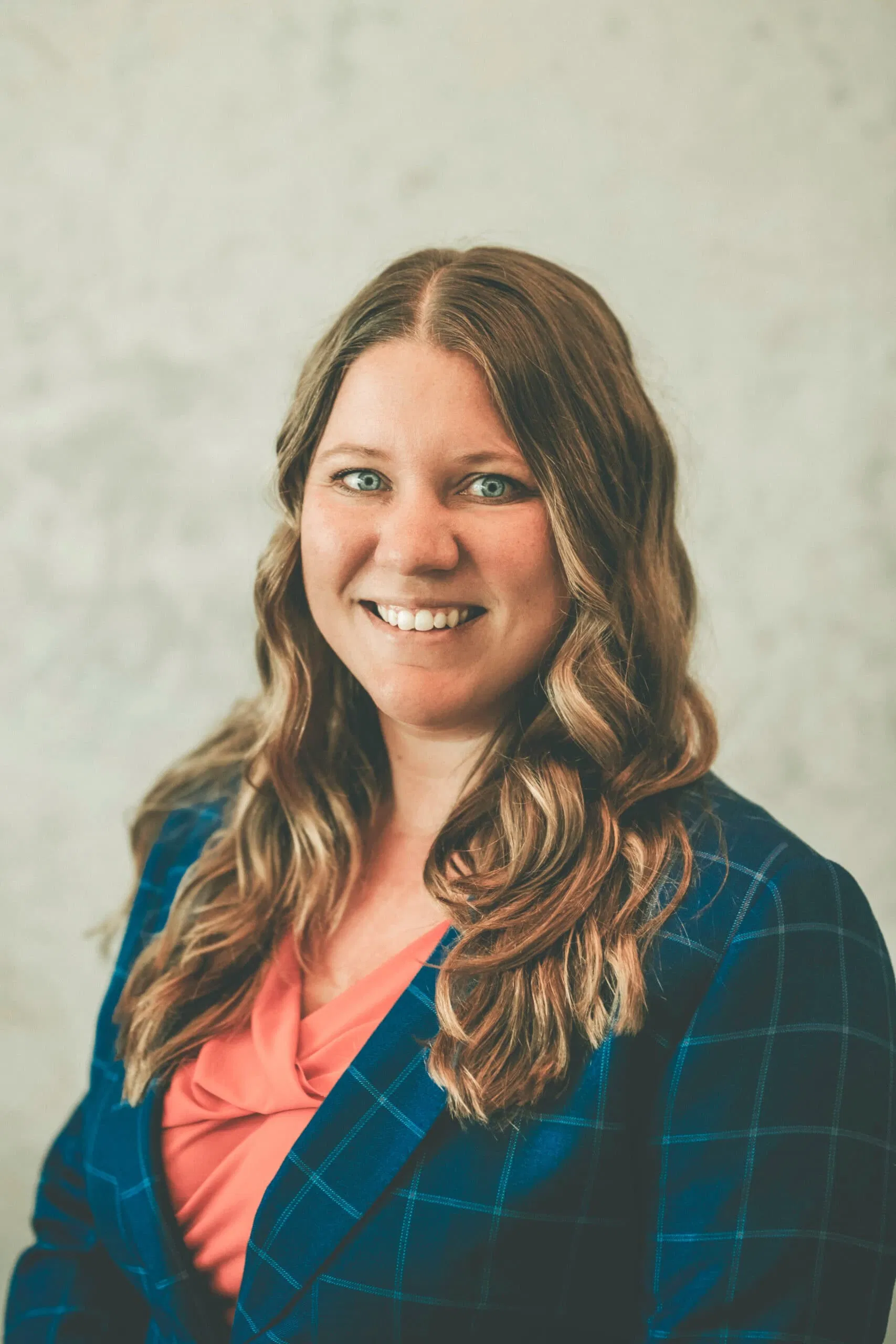 A person with long wavy hair, wearing a blue plaid blazer over an orange shirt, smiles at the camera against a neutral background. - Market Design Team: Define. Structure. Expand.
