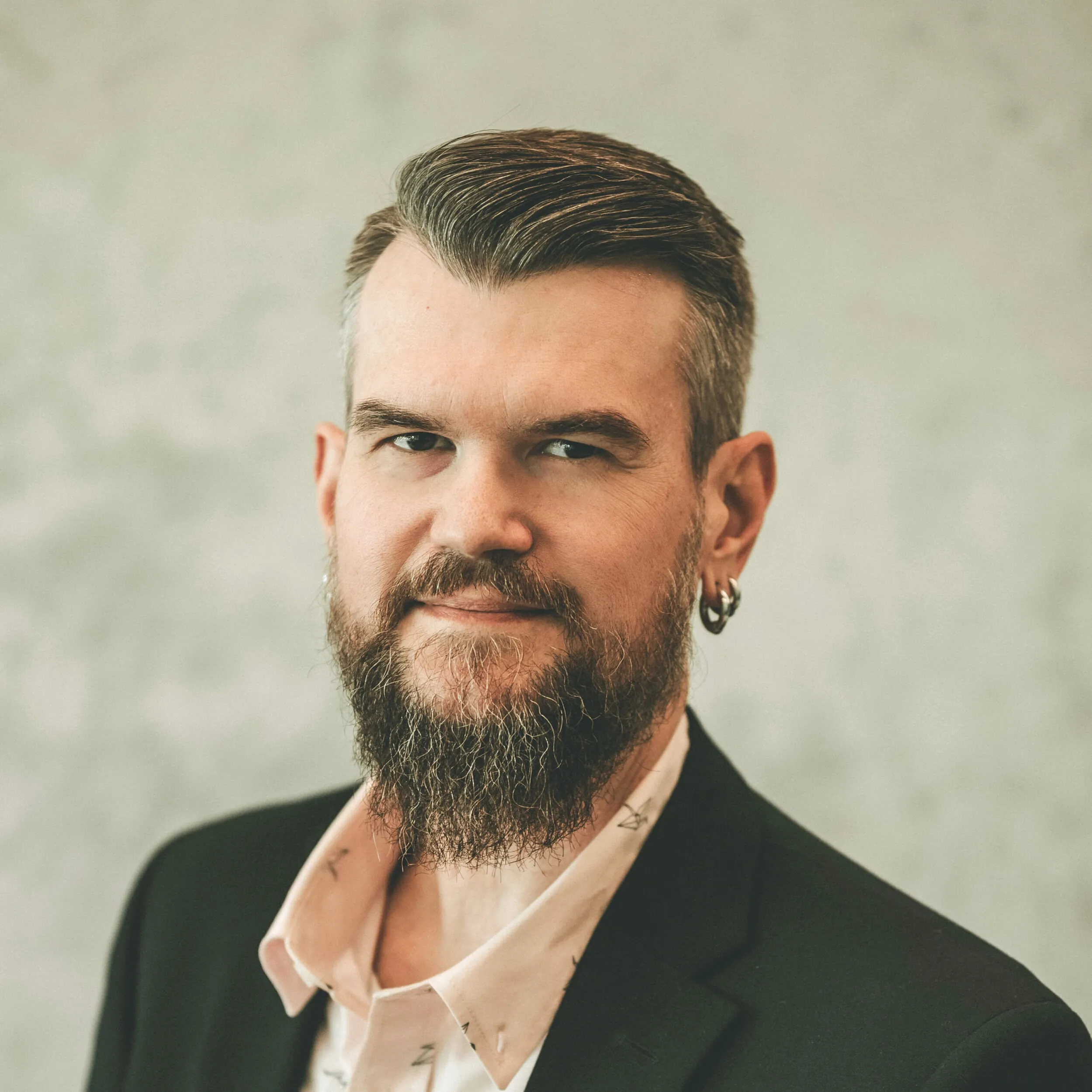 Daniel, sporting a beard and slicked-back hair, dons a black blazer over a white shirt. He has earrings in both ears and gazes slightly to the side. The neutral backdrop emphasizes his poised demeanor, perfect for those looking to schedule a meeting with him. - Market Design Team: Define. Structure. Expand.