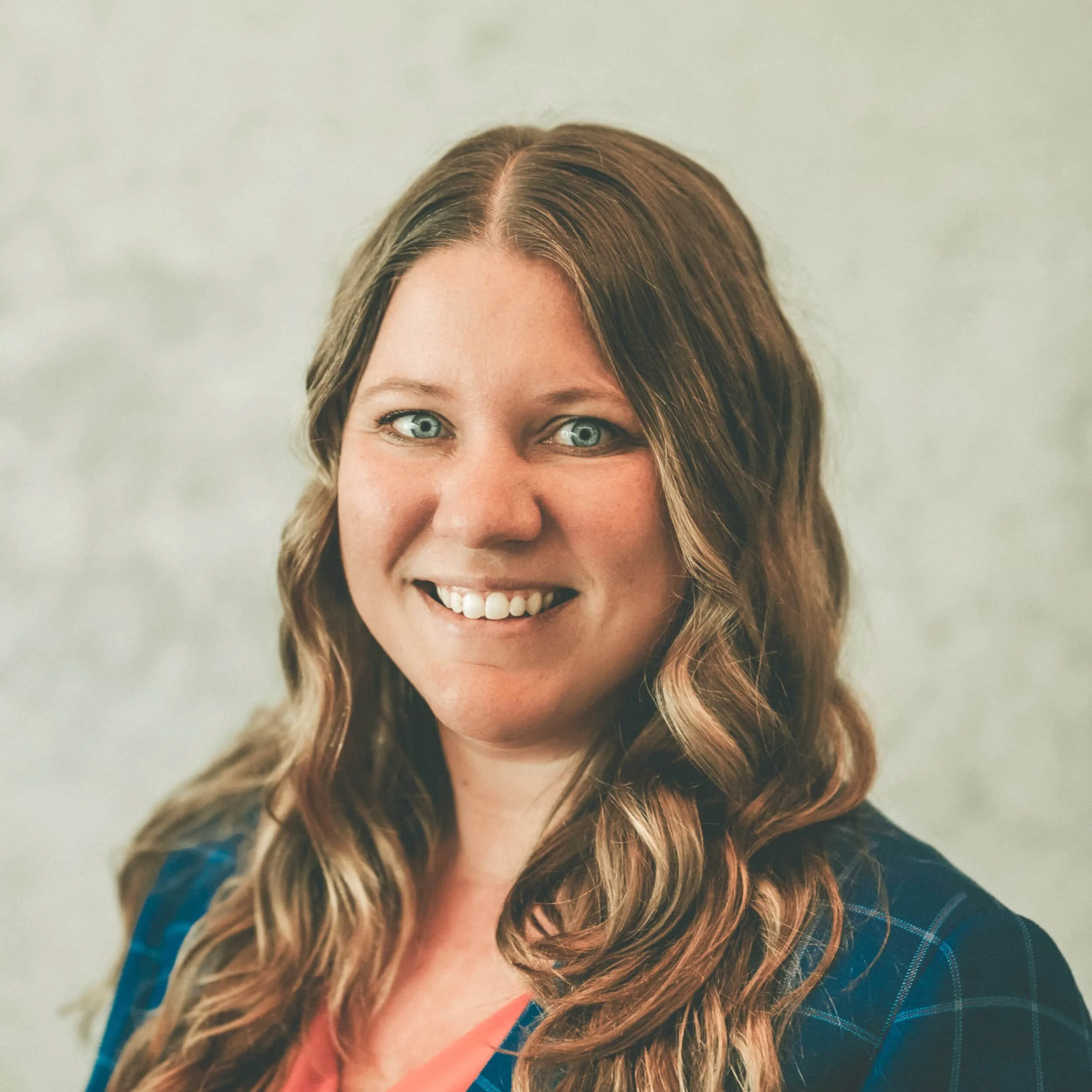 Margarey, with her long wavy hair and a radiant smile, dons a blue plaid jacket over a coral top as she faces the camera against a plain, light-colored background. - Market Design Team: Define. Structure. Expand.