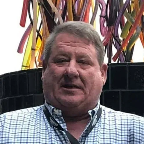 A man with short gray hair wearing a blue and white checkered shirt stands in front of a black Felder Water Well structure, with colorful vertical tubes or pipes in the background. - Market Design Team: Define. Structure. Expand.