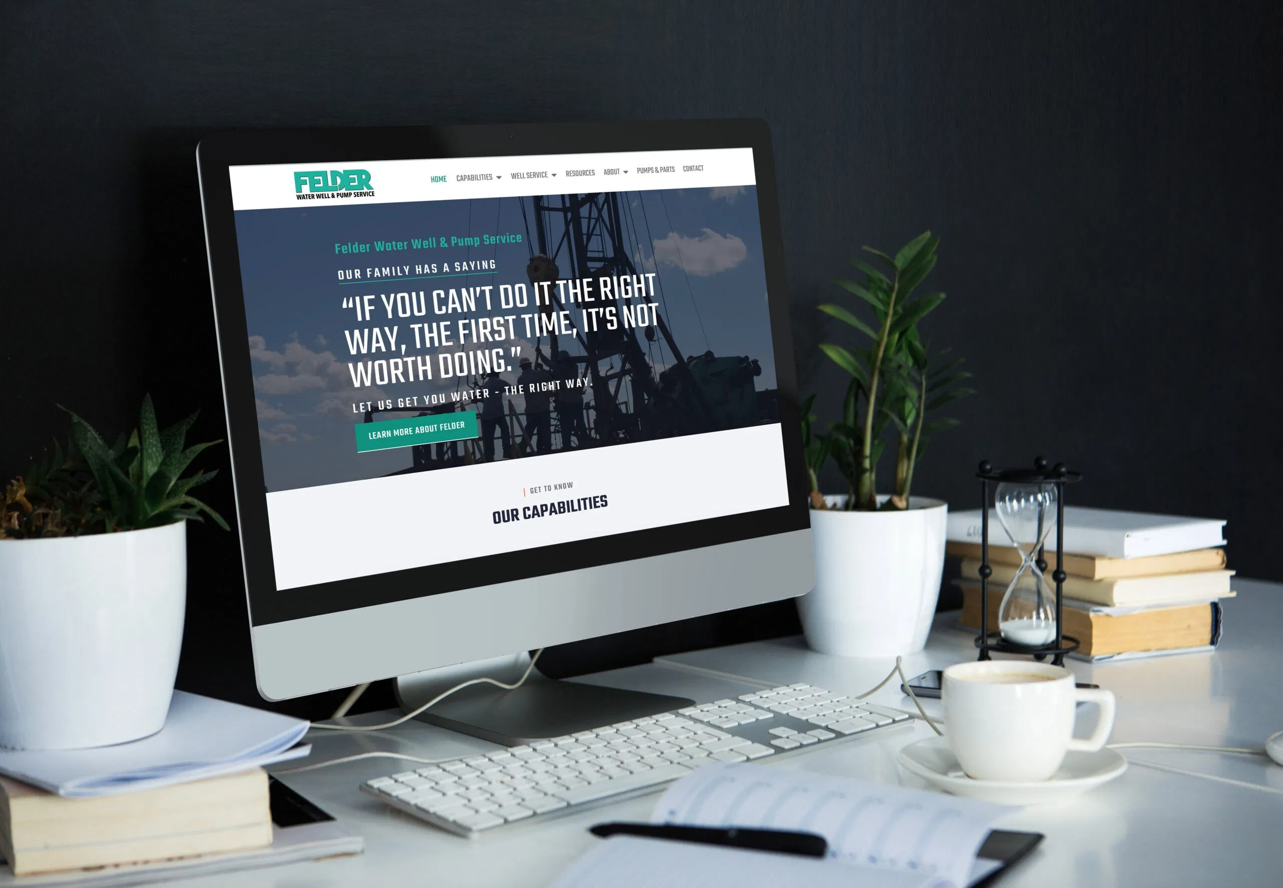 A desktop computer on a white desk displays the Felder Water Well website, featuring a large quote and navigation menu. The desk is neatly arranged with plants, a notebook, glasses, a pen, and a glass of water. - Market Design Team: Define. Structure. Expand.