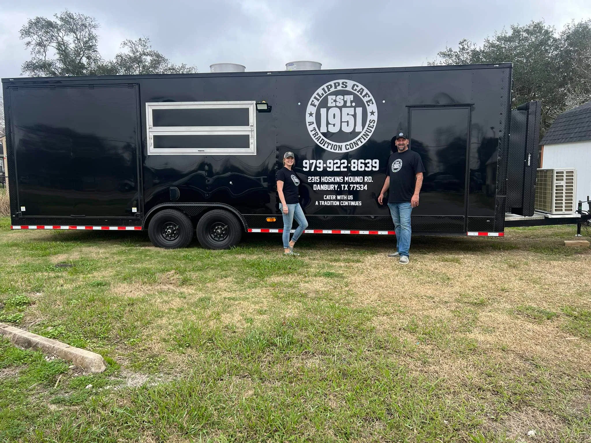 Two people stand in front of a large black Filipp's Cafe food trailer parked on grass. The trailer displays 