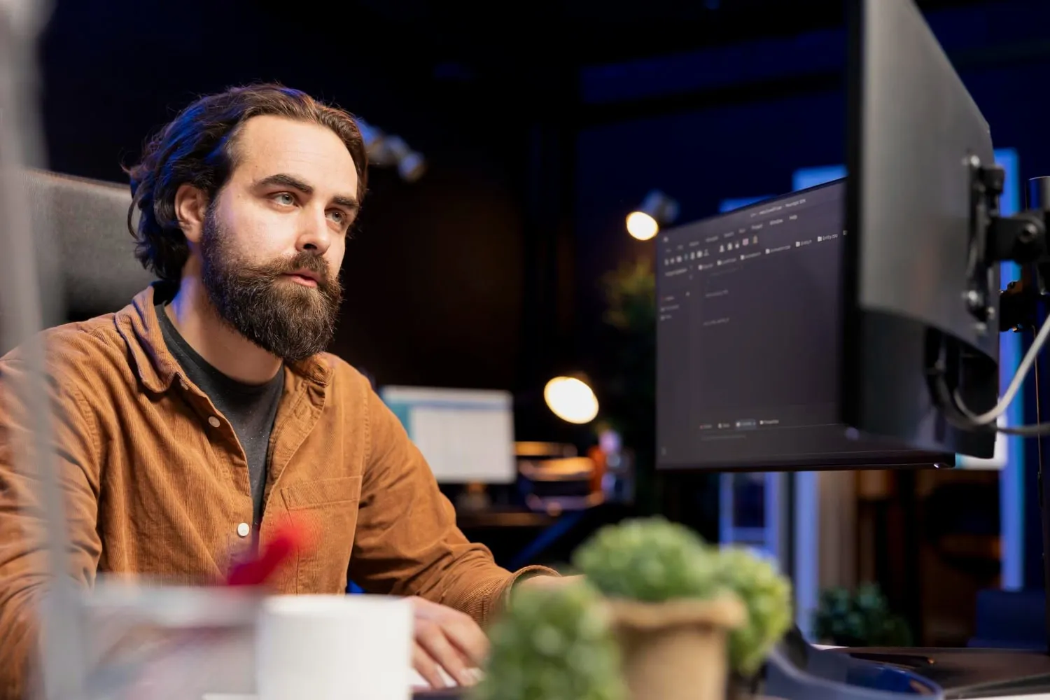 A man with a beard and long hair, wearing a brown shirt, sits at a desk and looks at a computer monitor in a dimly lit office with plants and equipment, focused on expert web development to streamline business operations. - Market Design Team: Define. Structure. Expand.