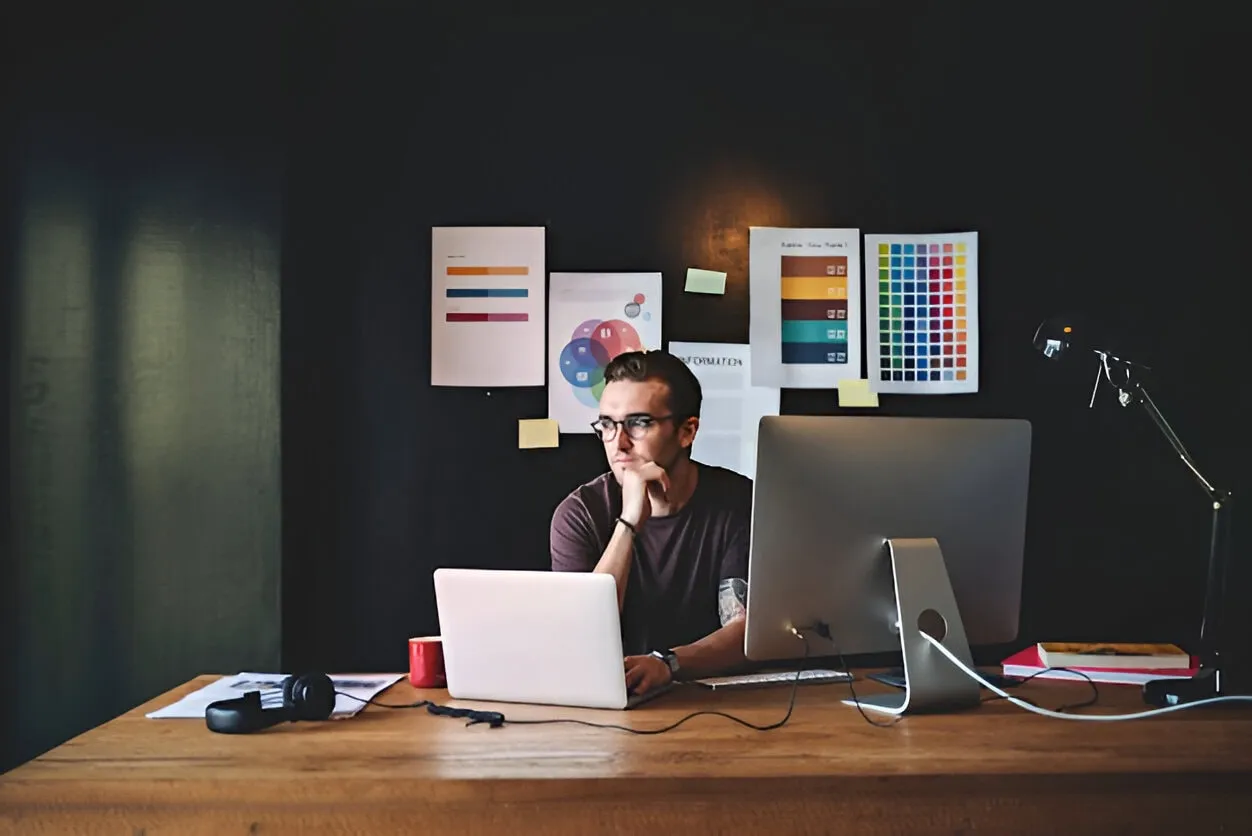 A web page designer sits at a desk with a laptop and desktop,