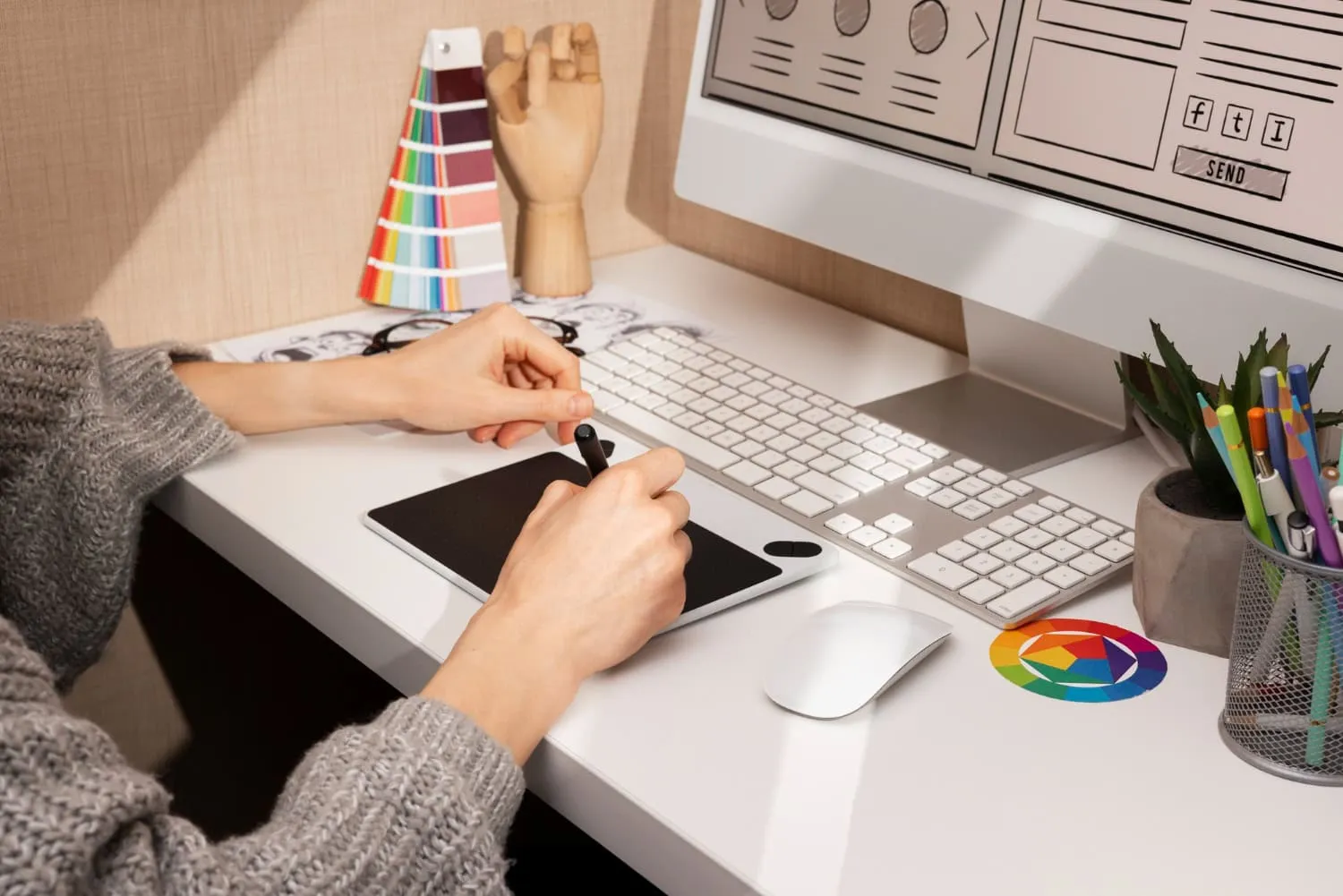 A person works on a digital drawing tablet at a white desk in Lake Jackson, focused on graphic design. A computer displays a design mockup, with color swatches, stationery, a wooden mannequin hand, and a color wheel coaster nearby. - Market Design Team: Define. Structure. Expand.
