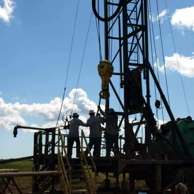 Three workers operate machinery on a Felder Water Well drilling rig outdoors during daylight, silhouetted against a blue sky with scattered clouds—a striking scene of well drilling in action. - Market Design Team: Define. Structure. Expand.