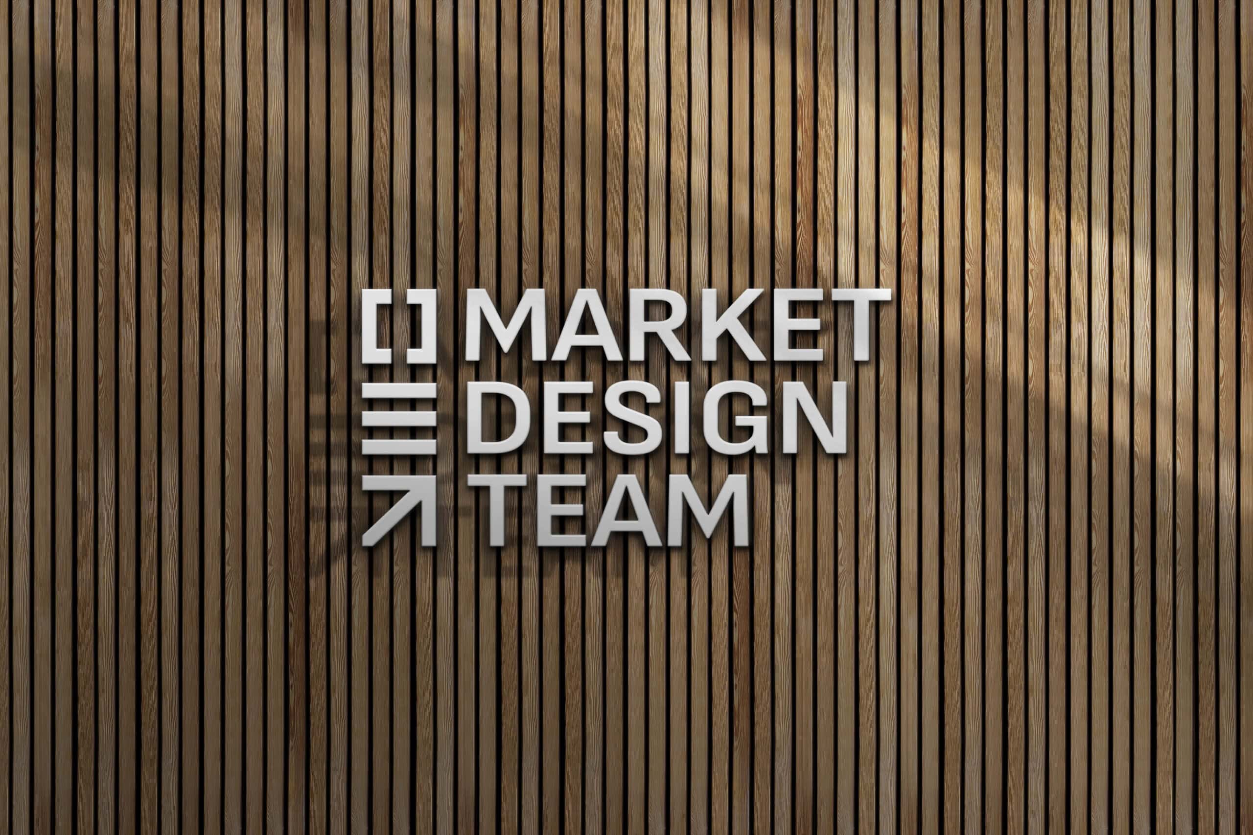 A vertical wooden plank wall serves as the background for a sign that reads "MARKET DESIGN TEAM" in modern, white, capital letters. The design includes horizontal, vertical, and diagonal line elements to the left of the text. - Market Design Team: Define. Structure. Expand.