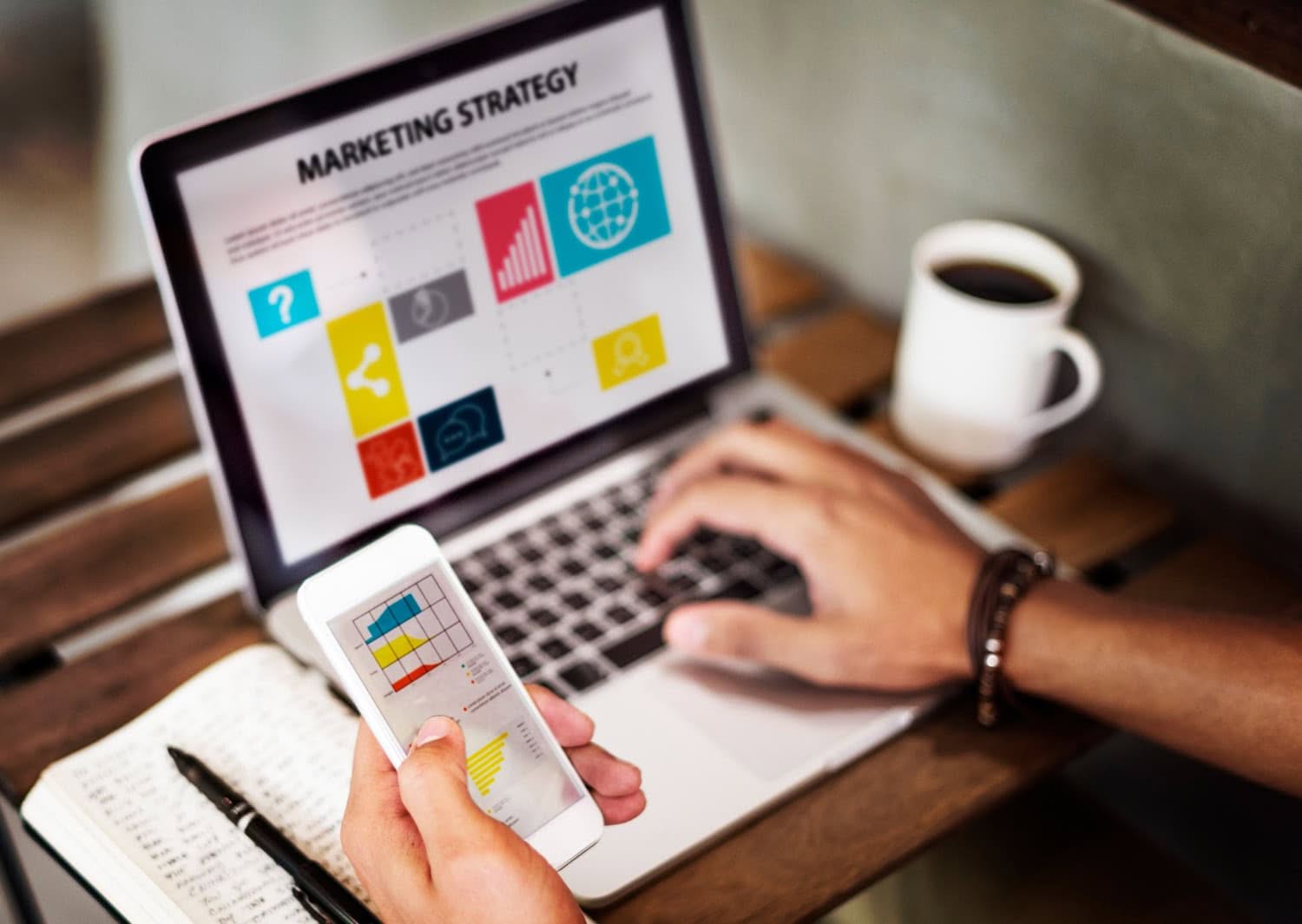 A person works on a laptop displaying a digital marketing strategy presentation with charts and icons, while holding a smartphone showing a graph. A notebook and cup of coffee sit on the wooden table, capturing a productive fall day in Lake Jackson. - Market Design Team: Define. Structure. Expand.
