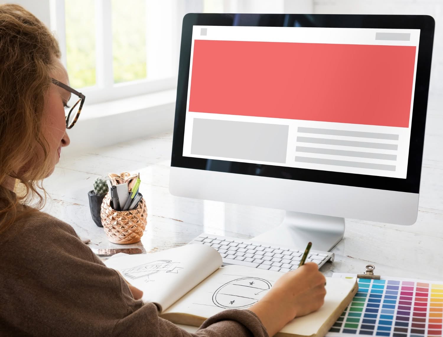 A person sits at a desk sketching in a notebook, with a color palette and computer monitor displaying a rural web design layout. Natural light streams through a nearby window, highlighting the creative process. - Market Design Team: Define. Structure. Expand.