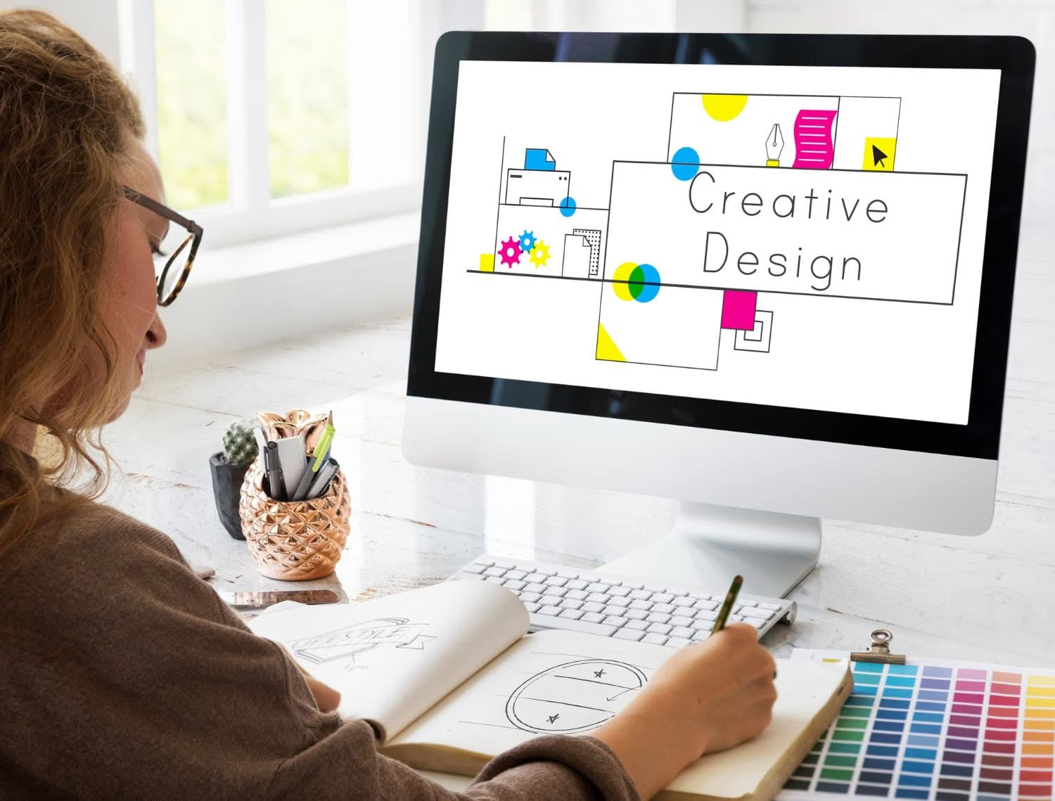 A person with long hair and glasses sketches in a notebook at a desk. A computer monitor displays a colorful design with the text "Creative Design." Art supplies and a plant are on the desk. - Market Design Team: Define. Structure. Expand.
